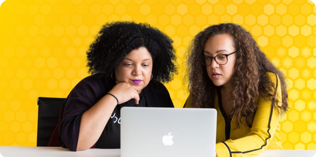 Two women sit side by side at a desk with a laptop, both are looking at the screen.
