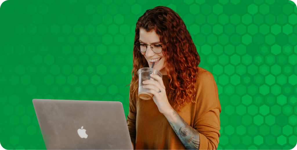 A DevOps engineer works at her laptop while smiling and drinking from an iced coffee with a straw.