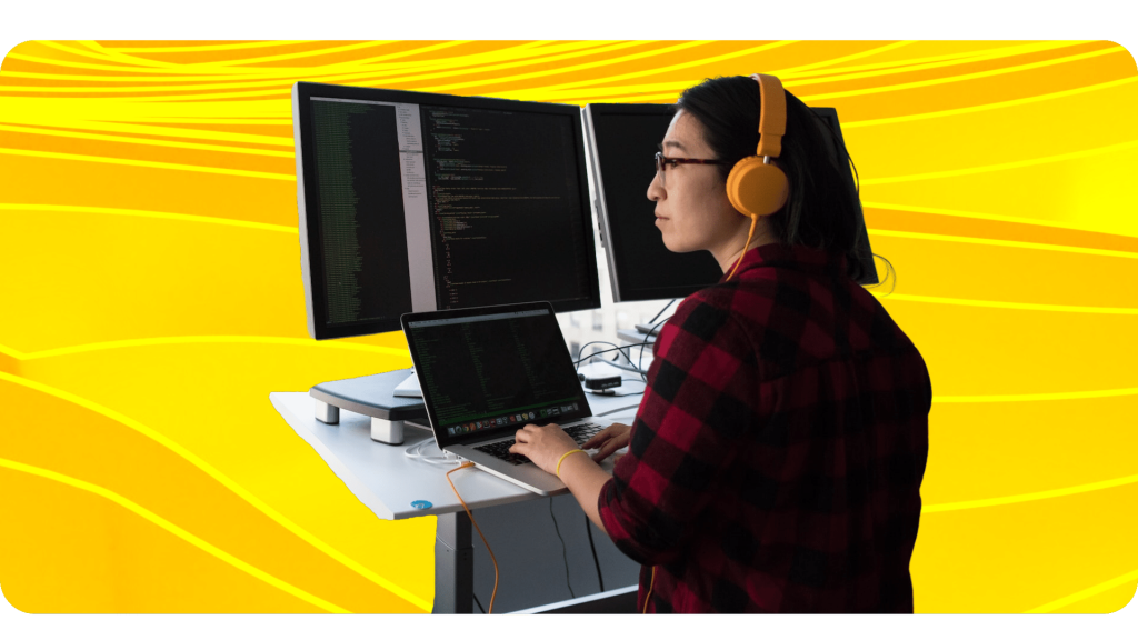 A software engineer types on a laptop while staring at lines of code on multiple monitors.