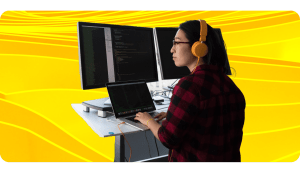 A software engineer types on a laptop while staring at lines of code on multiple monitors.