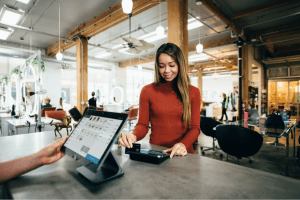 A woman swiping a card on a card reader inside a hair salon while a receptionist processes the transaction on a POS workstation.
