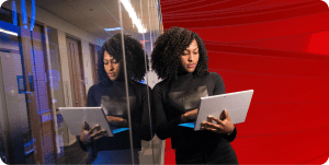 Woman leans against a glass wall holding a laptop.