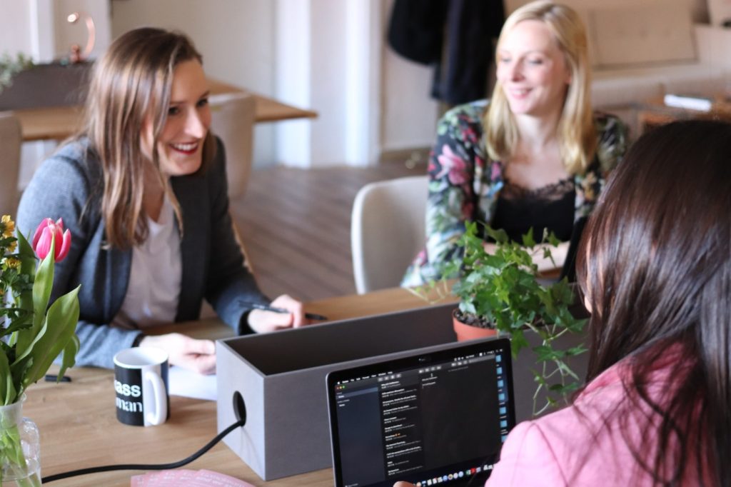 Three women sit around a desk working, the woman in the foreground has a laptop open.