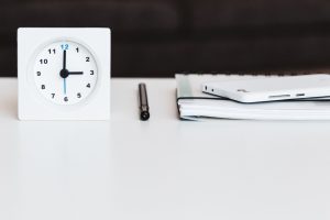 Square white clock on a white table next to a pen, notebook, and smartphone, on a black background.