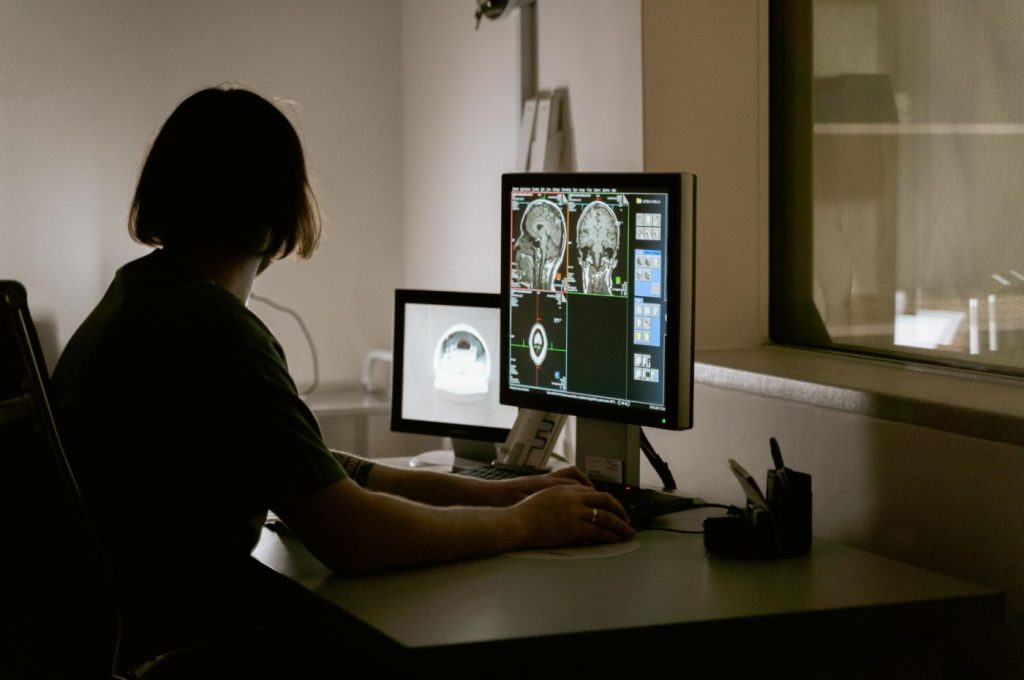 An MRI technician sits in front of a set of computer monitors, watching as diagnostic scans appear on-screen.