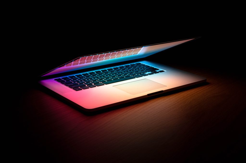 A half-closed MacBook laptop lights up its darkened surroundings in soft rainbow colors.