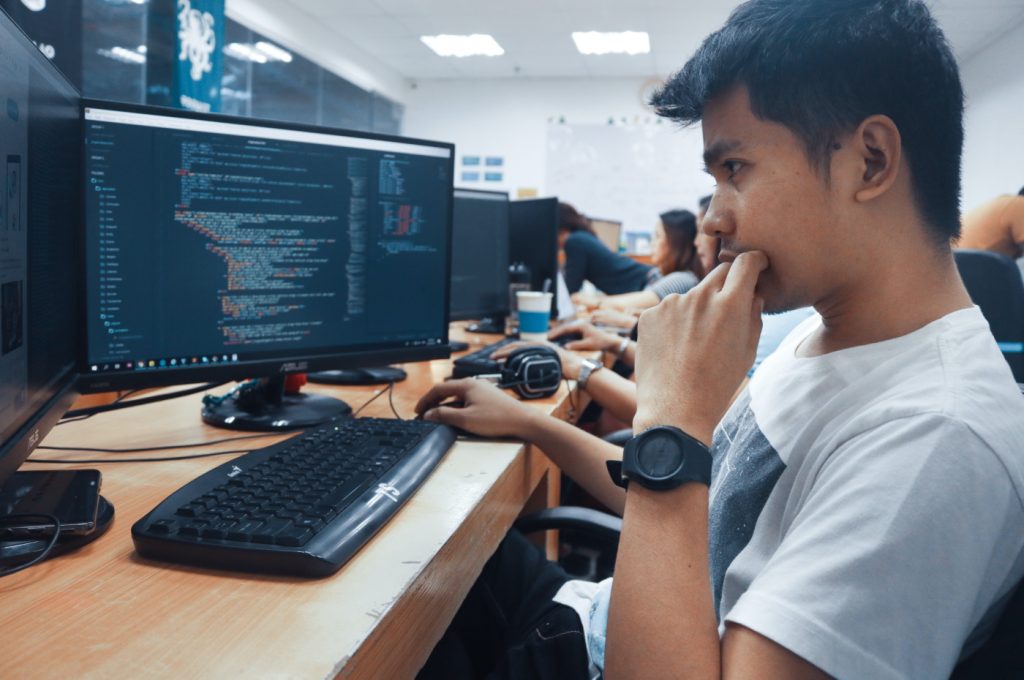 male engineer sitting in front of a computer screen in an office, studying lines of code