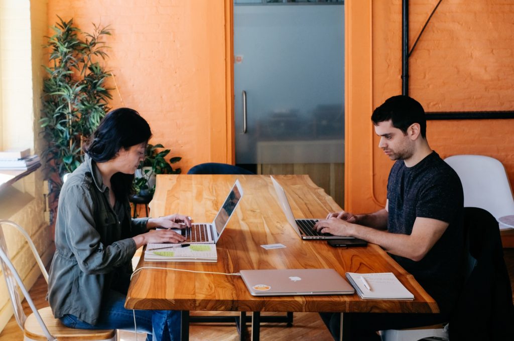 Two people working on laptops at a conference table in a brightly-lit office with orange walls.