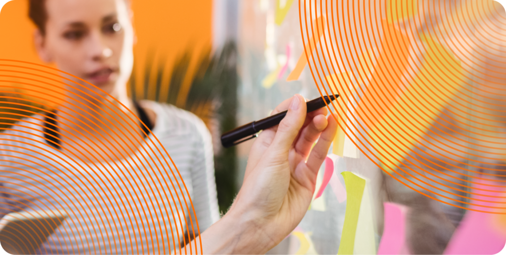 A close up of a hand writing on a sticky note attached to a clear wall, surrounded by other such sticky notes. In the background, a coworker looks on with interest.