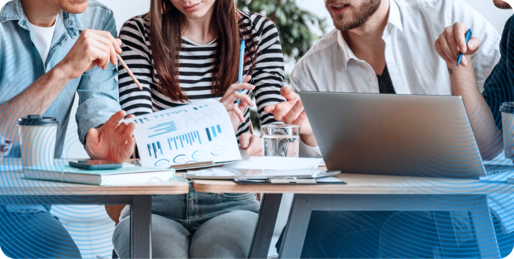 A close up of four coworkers collaborating and discussing work. One holds a clipboard full of printed reports, two more lean in to offer their opinions, and another takes notes using their laptop.