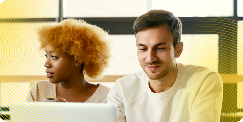 Two coworkers enjoy a bright and easy day at the office. One looks down at his laptop and smiles, while the other looks off-screen, holding a mug of coffee.