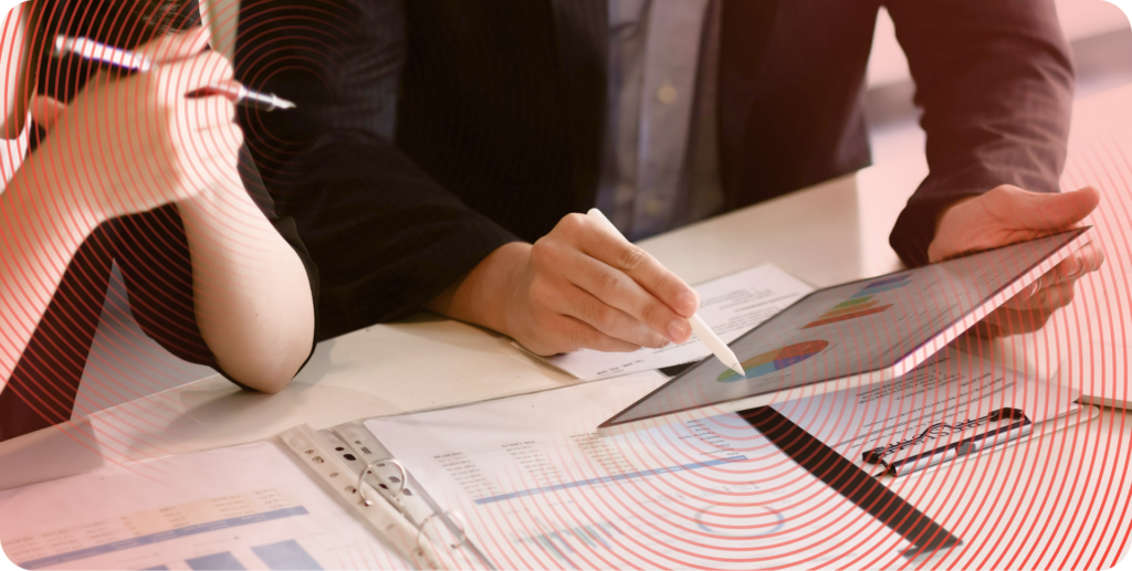 A close up of coworkers' hands while they discuss a report. One worker holds a tablet and points to a graph using a stylus, while the other rests their elbows on the edge of a table. A binder full of printed reports rests on the table.