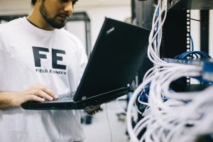 A man wearing a white shirt with the words "FE: Field Engineer" written in black holds a black laptop in a server room.