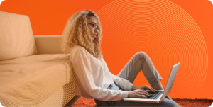 A woman with long blonde hair and glasses works on her laptop while seated on the floor, he back reclining against a couch.