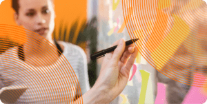 A close up of a hand writing on a sticky note attached to a clear wall, surrounded by other such sticky notes. In the background, a coworker looks on with interest.