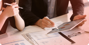 A close up of coworkers' hands while they discuss a report. One worker holds a tablet and points to a graph using a stylus, while the other rests their elbows on the edge of a table. A binder full of printed reports rests on the table.