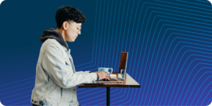 A person with short hair, glasses, and a denim jacket working on a laptop at a high table with a blue background featuring subtle geometric lines.