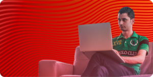 A person in a green and black polo shirt sitting on a couch and working on a laptop. The background is red with wavy horizontal lines, highlighting focus and productivity.