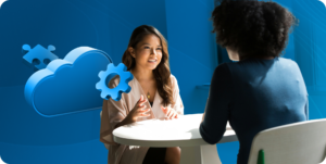 Two people seated at a round table having a discussion, with blue cloud and gear icons in the background.